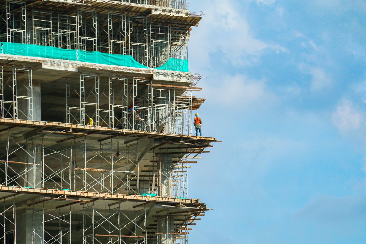 Home A construction worker stands on a scaffolding at a high-rise building under a blue sky.